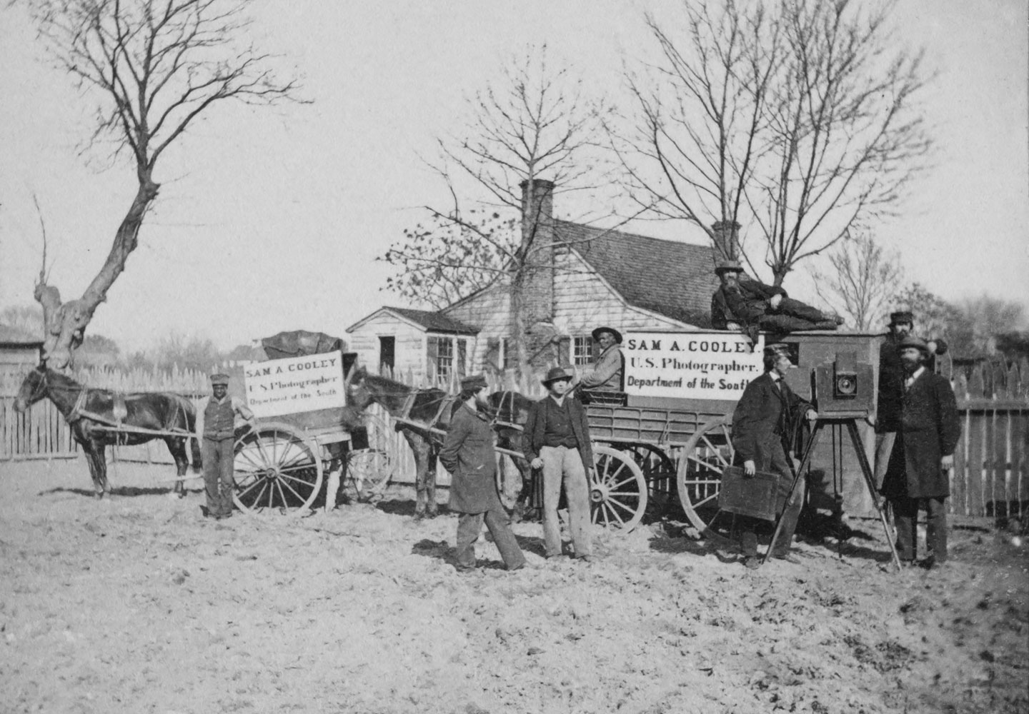 Stereograph shows a group of men standing next to wagons labeled 'Sam A. Cooley U.S. Photographer Department of the South', with a camera on the left, and two African American men employed as drivers
