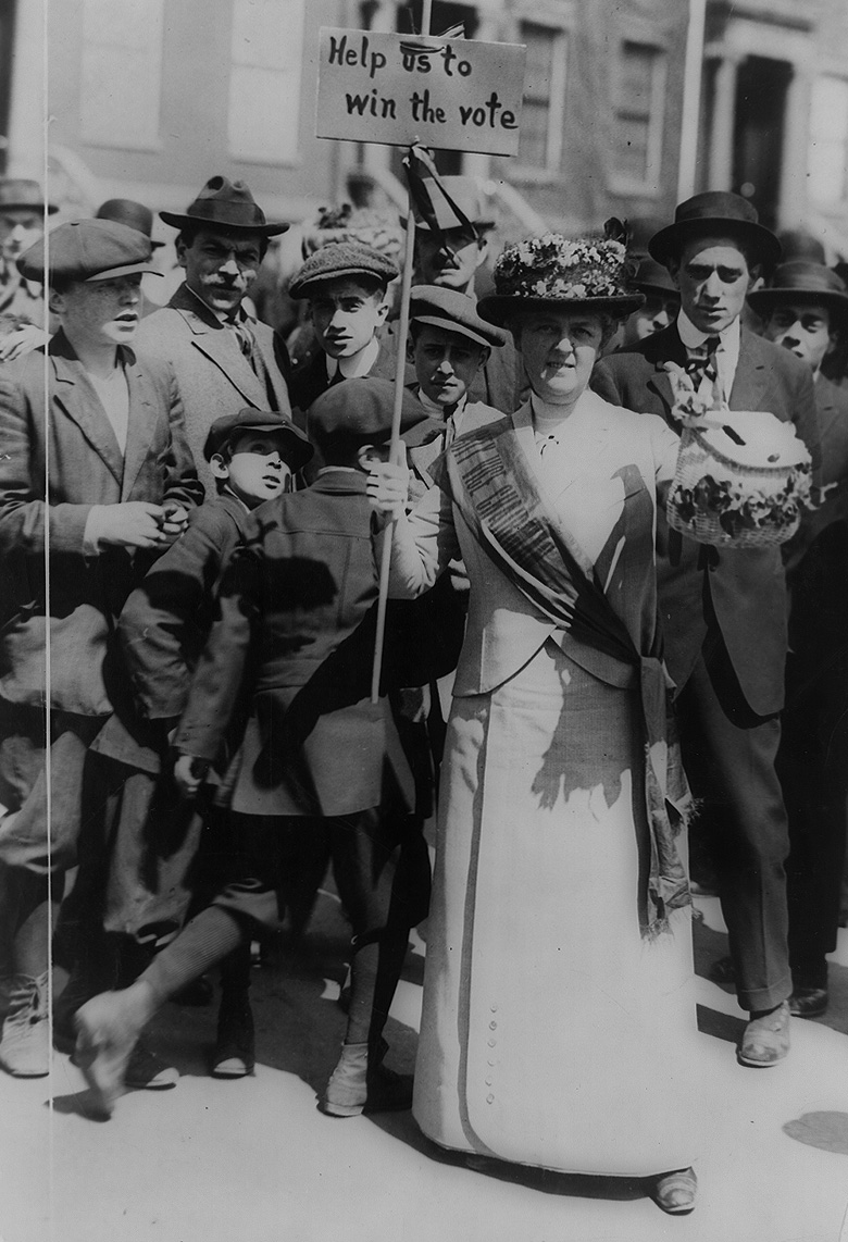 Suffragist, “Mrs. Suffern,” holding a sign and a flowered ballot box, amid a crowd of boys and men, 1914.